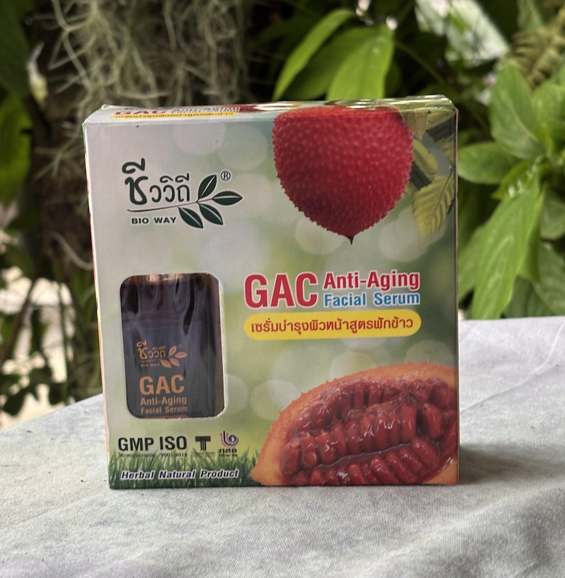 K&ouml;nnte beinhalten: Eine quadratische Pappschachtel f&uuml;r das Bio Way GAC Anti-Aging Gesichtsserum. Die Verpackung zeigt eine rote, stachelige Gac-Frucht, einen Querschnitt der Frucht und ein Sichtfenster mit einer dunkelbraunen Flasche. Der Text umfasst GAC Anti-Aging Facial Serum, GMP ISO und Herbal Natural Product.