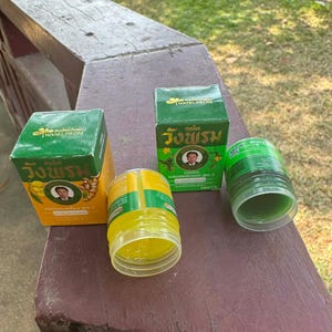 Pode incluir: Uma variedade de produtos de bálsamo de ervas Wang Prom. A imagem mostra dois potes de bálsamo verdes e amarelos, um aberto, e duas caixas verdes e amarelas. As caixas e os potes têm texto e um retrato de um homem.