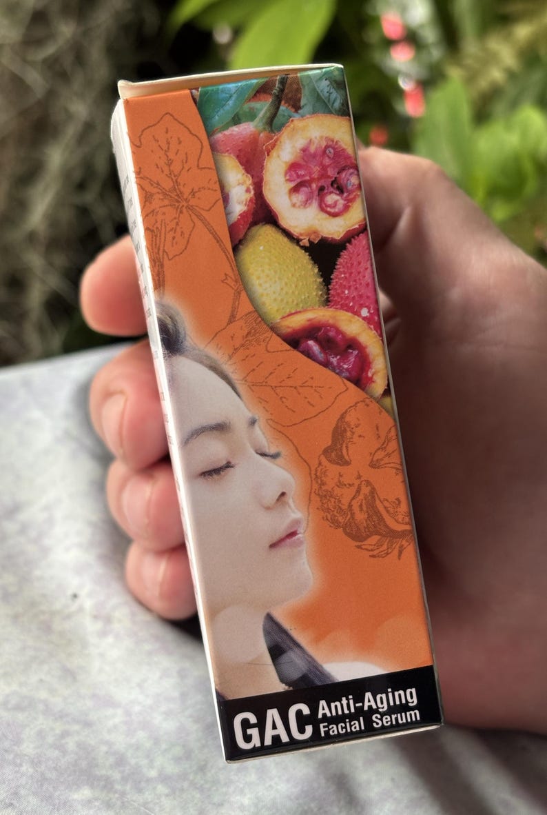 K&ouml;nnte beinhalten: Eine rechteckige Schachtel f&uuml;r GAC Anti-Aging Facial Serum mit orangefarbenem Hintergrund, Strichzeichnungen von Bl&auml;ttern, einem Foto von aufgeschnittenen Gac-Fr&uuml;chten und dem Profil einer Person mit geschlossenen Augen. Der Text GAC Anti-Aging Facial Serum ist aufgedruckt.