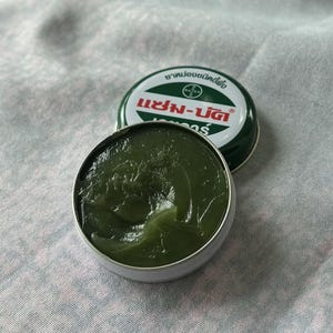 May include: Open tin of green ointment with the lid slightly ajar. The lid is green with white text and a red Bayer logo. The ointment is a dark green colour and has a smooth, glossy texture. The tin is silver.