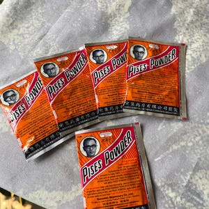 May include: Several orange and white packets of PISES POWDER are arranged on a patterned surface. Each packet features a portrait of a man in glasses and the product name in bold letters. The packets are sealed.