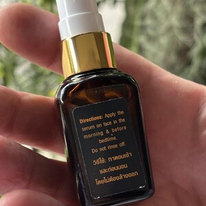 K&ouml;nnte beinhalten: Eine kleine bernsteinfarbene Glasflasche mit wei&szlig;em Pumpspender und goldenem Kragen. Ein schwarzes rechteckiges Etikett auf der Vorderseite zeigt wei&szlig;en und orangefarbenen Text mit der Anweisung: Apply the serum on face in the morning & before bedtime.