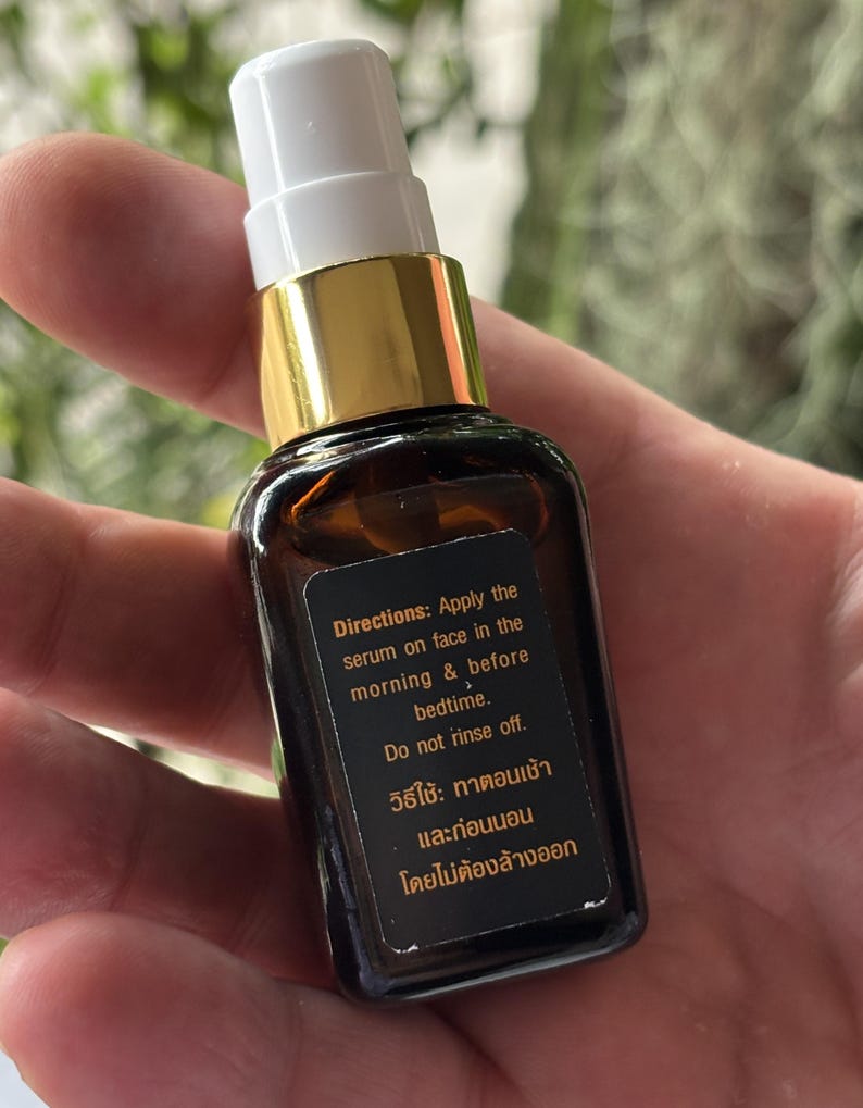 K&ouml;nnte beinhalten: Eine kleine bernsteinfarbene Glasflasche mit wei&szlig;em Pumpspender und goldenem Kragen. Ein schwarzes rechteckiges Etikett auf der Vorderseite zeigt wei&szlig;en und orangefarbenen Text mit der Anweisung: Apply the serum on face in the morning & before bedtime.