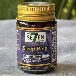 Op de afbeelding: Een klein glazen potje met een gouden deksel, met het opschrift "Sleep Balm" met de tekst "Lavender" en "Moisturizing Herb". Het etiket is paars en wit met groene accenten. Het potje bevat een donkerbruine substantie.