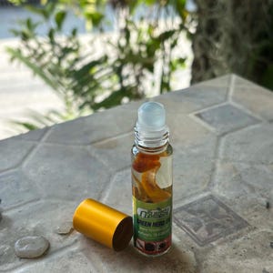 Puede incluir: Un frasco roll-on de vidrio transparente con una tapa dorada, que contiene un líquido con trozos de color naranja. El frasco tiene una etiqueta con texto verde que dice "GREEN HERB OIL". La tapa está al lado del frasco sobre una superficie de piedra.