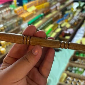 May include: A wooden tool with a cylindrical body and rounded ends. The tool has carved rings and a tapered end. The wood has a natural grain and a warm, brown colour. The tool is held in a hand.