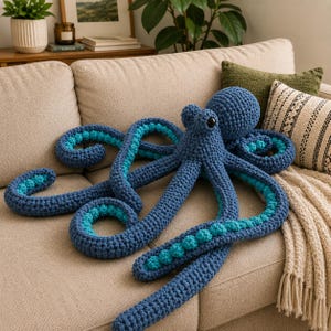 Puede incluir: Un gran pulpo azul tejido a mano con detalles turquesa en sus tentáculos descansa sobre un sofá beige. El pulpo tiene una cabeza redonda y ocho tentáculos largos y curvos. Los tentáculos presentan rayas y bolitas turquesas.