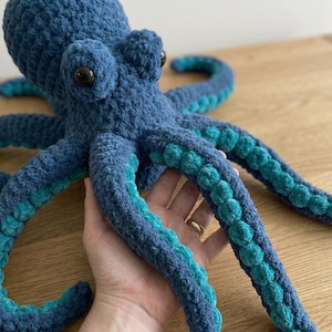 Könnte beinhalten: Handgefertigter Plüsch-Oktopus in Blau- und Türkistönen. Der Oktopus hat große, schwarze Augen und detaillierte Tentakel. Das Plüschtier besteht aus weichem, strukturiertem Garn, ideal für ein Kinderzimmer oder eine Dekoration im maritimen Stil.