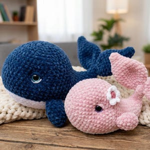 May include: Two plush crochet whales. One is navy blue with a white belly, and the other is pink with a white flower accent. Both have dark eyes and are resting on a cream-colored blanket. The whales are on a wooden surface.