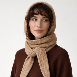 May include: A beige knitted hood and scarf set. The hood frames the face, and the scarf is wrapped around the neck and tied in front. The set is made of a thick, textured knit material. The model is wearing a brown sweater.