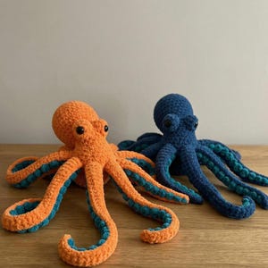 May include: Two crocheted octopus plush toys. One is orange with teal accents on its tentacles, and the other is blue with teal accents. Both have black eyes and are displayed on a wooden surface.
