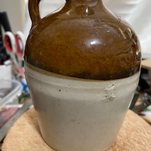 May include: A vintage stoneware jug with a handle. The upper portion is a rich brown, while the lower half is a creamy white. The jug has a rounded body and a narrow neck, suggesting it was used for storing liquids.