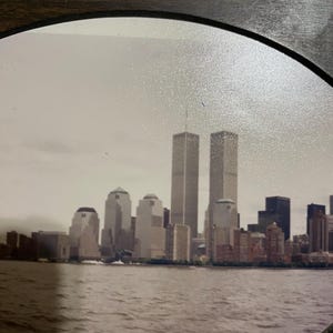 May include: A framed photograph of the New York City skyline, featuring the Twin Towers and other buildings along the waterfront. The image is in sepia tones, with the water in the foreground and a cloudy sky.