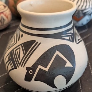 May include: A small, off-white ceramic vase with a rounded body and a wide opening. The vase is decorated with black geometric patterns and a stylized bear figure. The design includes lines, triangles, and a bear silhouette, creating a Southwestern aesthetic.