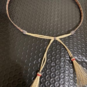 Op de afbeelding: Een cirkelvormige gevlochten koordriem met een dunne rode streep in het midden. De riem bevat verschillende kleine, donkere, getextureerde cilindrische kralen en eindigt in twee lange, lichtgekleurde vezelkwastjes met rode en donkere kraalaccenten.