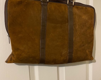 May include: A rectangular brown suede briefcase featuring a dark brown leather trim, two vertical leather straps, and a matching brown handle. The bag includes a top zipper closure with a metal pull and is suspended from a clear plastic hanger.