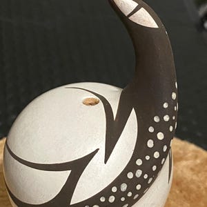 May include: A spherical ceramic shaker with a matte white finish and a curved, dark brown neck. The dark brown section features a pattern of white dots and a single white eye-like shape. A small circular opening with a cork stopper is visible.
