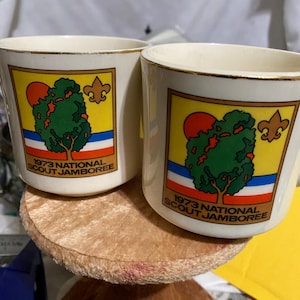 Könnte beinhalten: Zwei cremefarbene Keramikbecher mit Goldrändern. Jeder Becher zeigt eine farbenfrohe Grafik eines Baumes, einer Sonne und eines Pfadfinder-Emblems auf gelbem Hintergrund mit dem Text "1973 National Scout Jamboree".