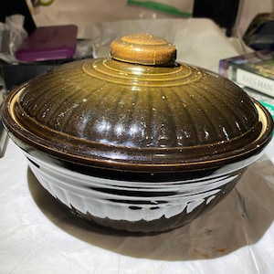 Puede incluir: Una fuente de cerámica marrón con tapa. El plato tiene forma redondeada con exterior marrón oscuro e interior marrón más claro. La tapa está rematada con un pomo de madera. El plato es probablemente vintage y apto para cocinar.