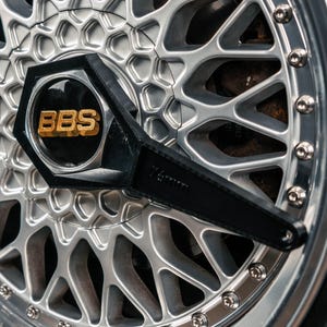 Könnte beinhalten: Nahaufnahme eines silber-schwarzen Autorads. Das Rad hat ein komplexes Speichendesign und eine glänzende, polierte Felge. Die Radkappe zeigt die goldenen Buchstaben "BBS" und eine schwarze sechseckige Abdeckung. Ein schwarzes Werkzeug ist am Rad befestigt.