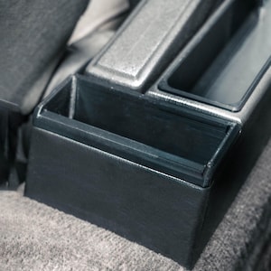 Könnte beinhalten: Nahaufnahme der Mittelkonsole eines Autos mit einem schwarzen, lederartigen Staufach und silberfarbenen Zierleisten. Die Konsole hat mehrere Bereiche, darunter ein kleines offenes Fach und einen größeren, abgedeckten Bereich. Das Design ist schlicht und modern.