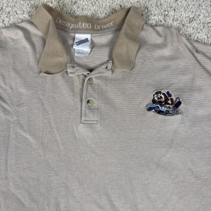Puede incluir: Un polo de color beige con cuello marrón claro que incluye el texto "Designated Driver" en la cinta interior del cuello. La prenda tiene una tapeta con dos botones y un bordado en el pecho que muestra al Demonio de Tasmania conduciendo.