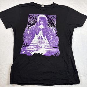 Puede incluir: Camiseta negra con un gráfico morado de la película Labyrinth. El gráfico incluye al Rey Goblin, un laberinto y otros personajes. La camiseta está hecha de un material suave.