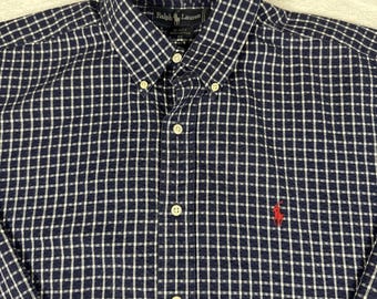 Puede incluir: Camisa abotonada a cuadros azul marino y blanco con cuello abotonado. La camisa presenta un pequeño logotipo rojo en el pecho y el nombre de la marca "Ralph Lauren" en la etiqueta.