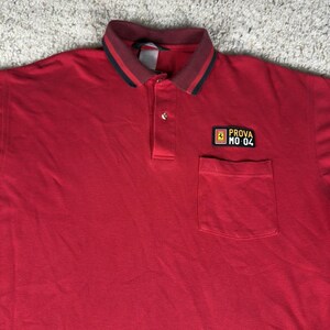May include: A red polo shirt with a collar featuring black and green stripes. The shirt has a pocket on the left chest and a small patch with the text "PROVA MO 04".