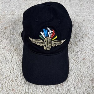 May include: A black baseball cap featuring an embroidered logo on the front. The logo displays a gold tire with wings, the text Indianapolis Motor Speedway, and a fan of colorful flags in white, blue, green, red, and a black and white checkered pattern.