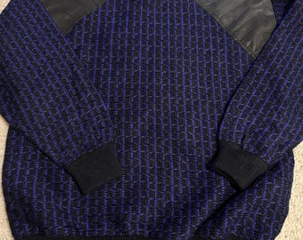 May include: A blue and black patterned sweater with black leather shoulder patches. The sweater has a ribbed collar, cuffs, and hem. The sleeves are long, and the overall style is vintage.