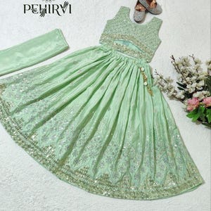 May include: A mint green two-piece outfit with intricate silver embroidery and sequin detailing. The set includes a crop top, a flowing skirt, and a matching scarf. The outfit is embellished with a gold and pearl tassel. The brand name "PEHIRVI" is visible.