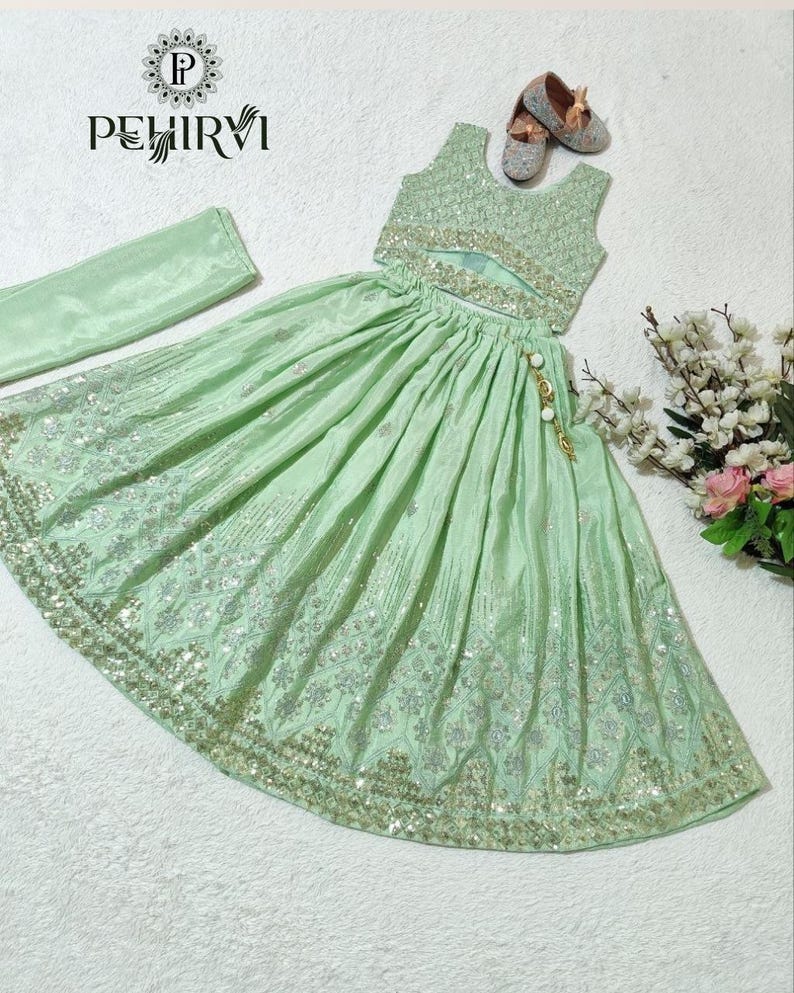 May include: A mint green two-piece outfit with intricate silver embroidery and sequin detailing. The set includes a crop top, a flowing skirt, and a matching scarf. The outfit is embellished with a gold and pearl tassel. The brand name "PEHIRVI" is visible.