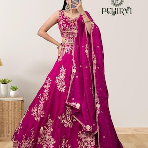 Puede incluir: Un vestido formal magenta sin mangas con intrincados bordados dorados. El vestido presenta una falda amplia y una bufanda transparente a juego. La parte superior está adornada con detalles dorados. El logotipo de la marca "PEHIRVI" es visible.