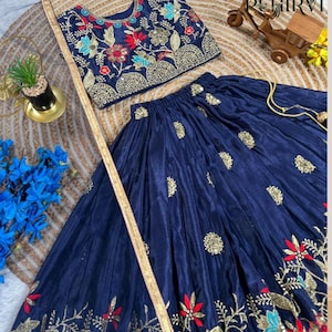 Op de afbeelding: Een marineblauwe tweedelige outfit met borduursels. De top is een mouwloze crop top met bloemenborduurwerk in rood, blauw en goud. De rok heeft een zwierig ontwerp met bijpassende borduursels. De outfit is gemaakt van een glanzende stof.