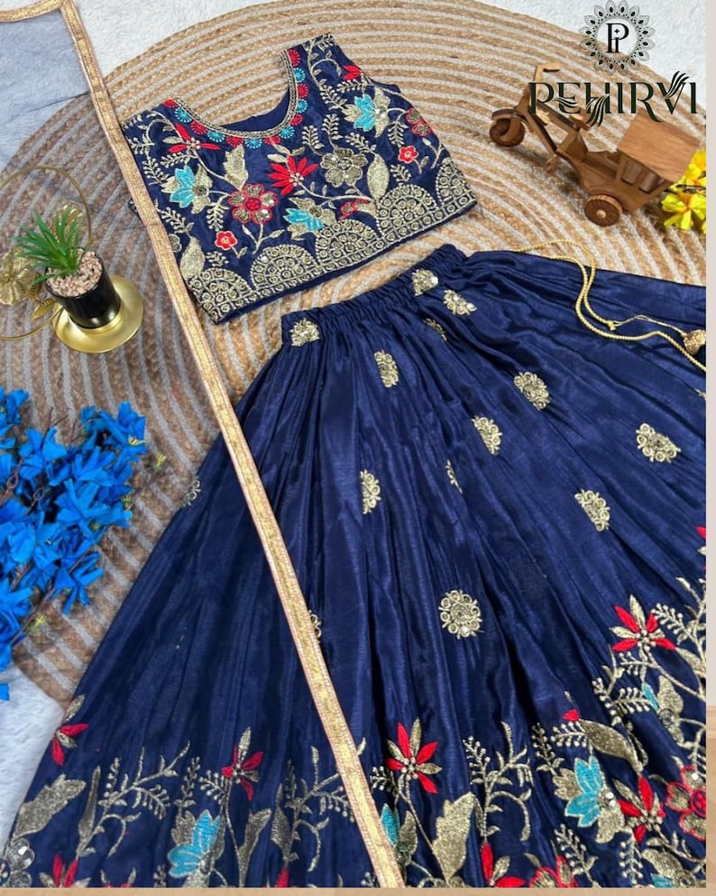 Op de afbeelding: Een marineblauwe tweedelige outfit met borduursels. De top is een mouwloze crop top met bloemenborduurwerk in rood, blauw en goud. De rok heeft een zwierig ontwerp met bijpassende borduursels. De outfit is gemaakt van een glanzende stof.