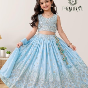 May include: A light blue, sleeveless crop top and skirt set with gold embellishments. The skirt has a flowing design with intricate detailing. The outfit includes a matching scarf and is accessorized with a beaded tassel. The brand logo "PEHIRVI" is visible.