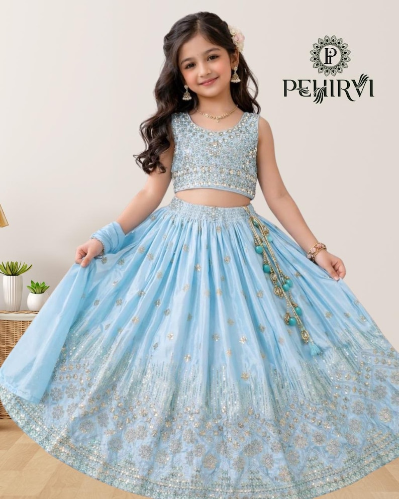 May include: A light blue, sleeveless crop top and skirt set with gold embellishments. The skirt has a flowing design with intricate detailing. The outfit includes a matching scarf and is accessorized with a beaded tassel. The brand logo "PEHIRVI" is visible.