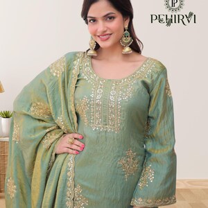 May include: A light green silk salwar kameez with gold embroidery. The outfit includes a long-sleeved top, matching pants, and a draped scarf. The top features intricate gold detailing around the neckline and cuffs. The image also shows gold earrings.