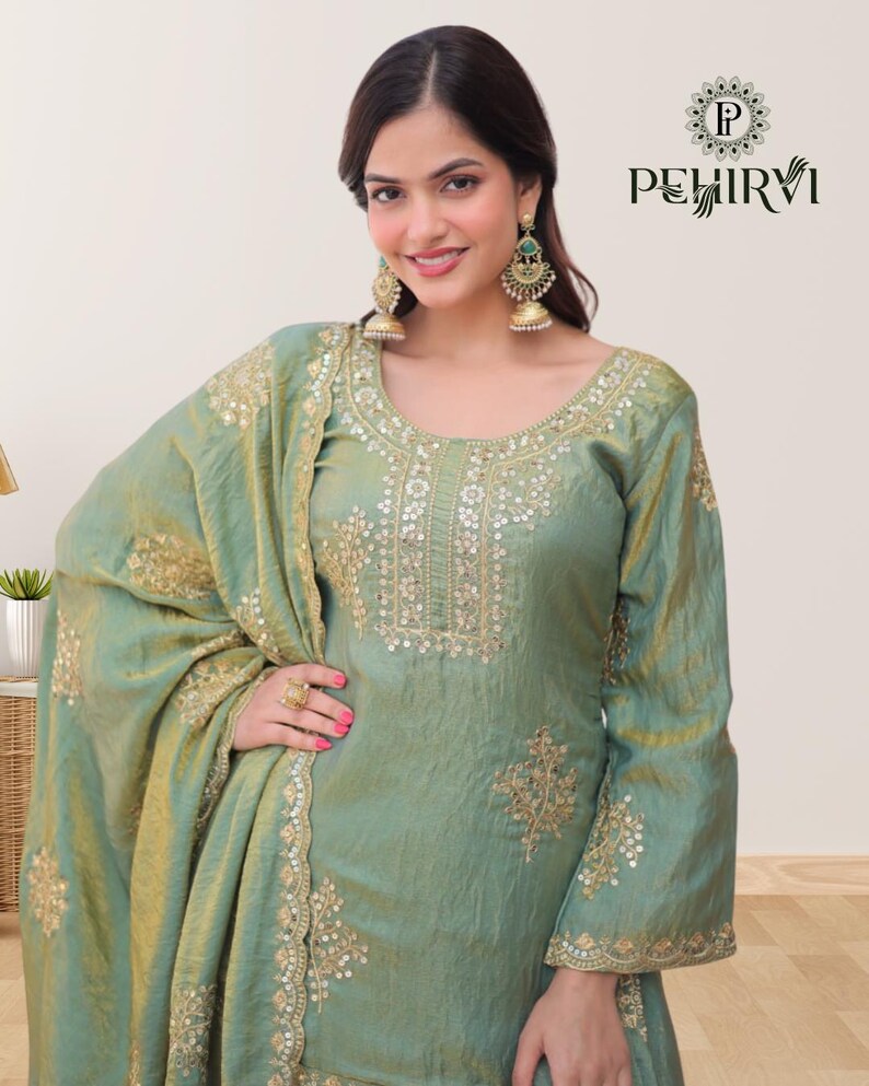 May include: A light green silk salwar kameez with gold embroidery. The outfit includes a long-sleeved top, matching pants, and a draped scarf. The top features intricate gold detailing around the neckline and cuffs. The image also shows gold earrings.
