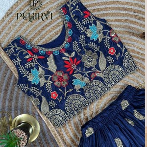 Op de afbeelding: Marineblauw geborduurde top en rok set. De top heeft ingewikkelde bloemenborduursels in rood, blauw en goud. De rok is marineblauw met gouden accenten. De set wordt gepresenteerd op een geweven achtergrond.