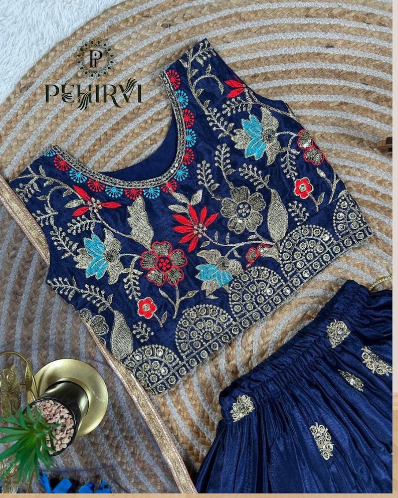 Op de afbeelding: Marineblauw geborduurde top en rok set. De top heeft ingewikkelde bloemenborduursels in rood, blauw en goud. De rok is marineblauw met gouden accenten. De set wordt gepresenteerd op een geweven achtergrond.