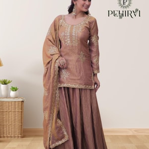 May include: A dusty rose-colored salwar kameez set. The long-sleeved top and wide-legged pants are embellished with gold embroidery. A matching dupatta is draped over the shoulder. The brand logo "PEHIRVI" is visible in the upper right corner.