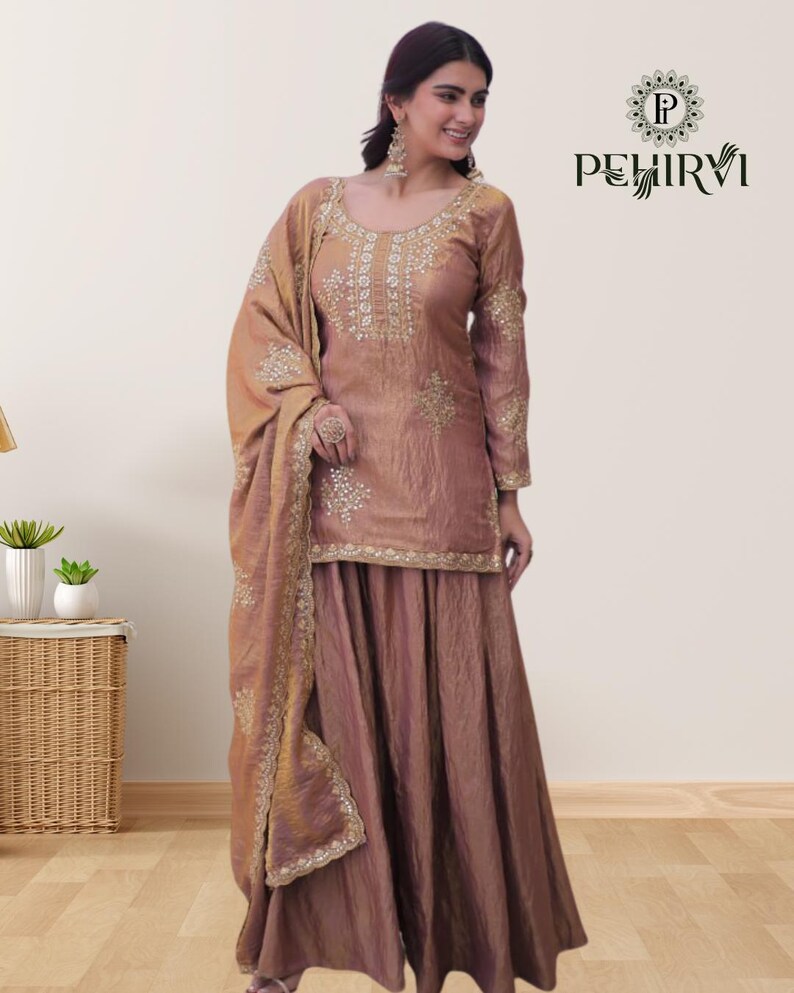 May include: A dusty rose-colored salwar kameez set. The long-sleeved top and wide-legged pants are embellished with gold embroidery. A matching dupatta is draped over the shoulder. The brand logo "PEHIRVI" is visible in the upper right corner.