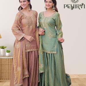 May include: Two women wearing matching traditional Indian outfits. The outfits feature long-sleeved tops and flowing skirts in shades of dusty rose and sage green. Intricate gold embroidery adorns the neckline, sleeves, and skirt hems. The outfits include matching scarves.