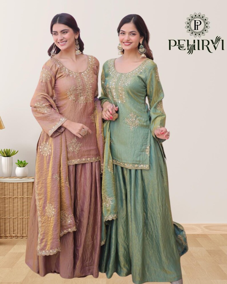 May include: Two women wearing matching traditional Indian outfits. The outfits feature long-sleeved tops and flowing skirts in shades of dusty rose and sage green. Intricate gold embroidery adorns the neckline, sleeves, and skirt hems. The outfits include matching scarves.