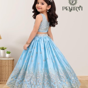 May include: A young girl wearing a light blue two-piece formal dress. The top is embellished with sequins, and the skirt features intricate gold embroidery. The dress is designed for special occasions.