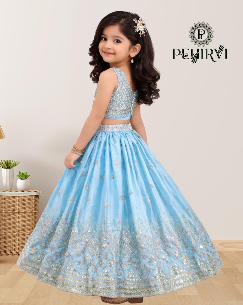 May include: A young girl wearing a light blue two-piece formal dress. The top is embellished with sequins, and the skirt features intricate gold embroidery. The dress is designed for special occasions.