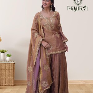 May include: A formal, two-piece outfit in shades of brown and purple. The top and dupatta feature intricate gold embroidery. The outfit is worn with matching wide-leg pants. The brand logo "PEHIRVI" is visible in the upper right corner.