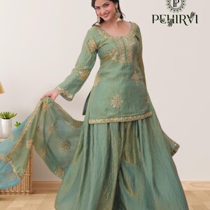 May include: A seafoam green Indian salwar kameez set with gold embroidery. The set includes a long-sleeved tunic, wide-legged pants, and a matching scarf. The tunic and pants are embellished with intricate gold detailing. The outfit is worn by a person.